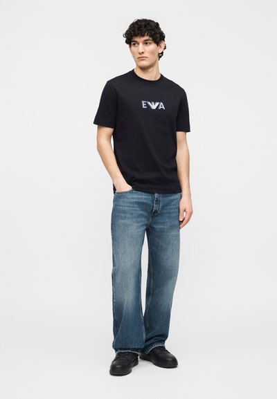 Νεαρός με σγουρά μαλλιά, φορώντας μαύρο T-shirt με λογότυπο EVA, μπλε τζιν παντελόνι φαρδιάς γραμμής και μαύρα αθλητικά παπούτσια, στέκεται με τα χέρια στις τσέπες.