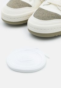 White sneakers feature a textured fabric upper with green accents, a rounded toe, and a chunky sole. A roll of white shoelaces is shown.