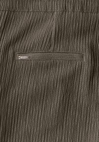 Textuur bruin stof met verticale ribbels. Bevat een klein, rechthoekig metalen label aan de linkerkant, met een verborgen zak.