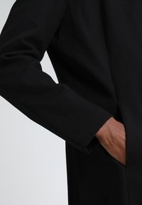 Abrigo negro con un tejido texturizado, silueta ajustada y un bolsillo lateral. La manga se extiende justo por encima de la muñeca.