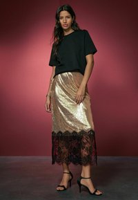 Schwarzes kurzärmliges Oberteil, kombiniert mit einem goldenen Paillettenrock mit schwarzem Spitzenrand. Schwarze High-Heels runden den Look ab.