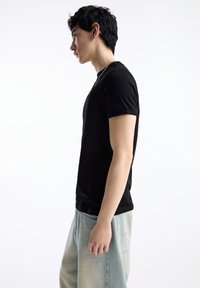 Tricou negru din bumbac, cu mâneci scurte, decolteu rotund, cu etichetă pe cusătură laterală, purtat cu blugi supradimensionați de culoare albastru deschis, văzut din lateral.