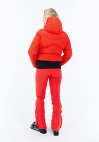 Mujer vestida con una chaqueta acolchada roja y pantalones de esquí rojos, con botas de esquí negras, de pie de espaldas sobre un fondo blanco.