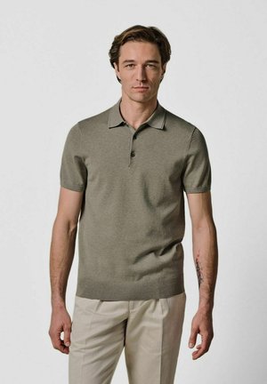 Polo de manga corta en oliva, de tejido suave, con tres botones, detalle en el cuello y diseño ajustado, combinado con pantalones de color claro.