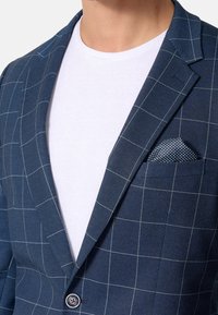 Chaqueta de traje azul marino con patrón de cuadros, solapa con muesca y pañuelo de bolsillo texturizado, combinada con una camiseta blanca lisa debajo.