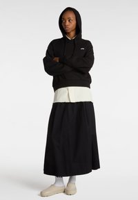 Sweatshirt noir à capuche avec poche avant, superposé sur une chemise blanche à rayures. Une jupe large noire et des chaussures beige à enfiler complètent le look.