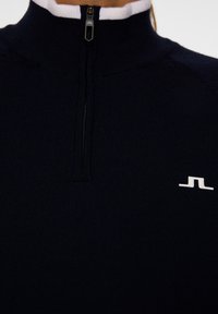 Laivastonsininen fleece-pulloväri, jossa on neljännesvetoketju, valkoinen kaulus, ribbatistruktuuri ja pieni valkoinen logo rinnassa. Yksinkertainen ja toiminnallinen muotoilu.