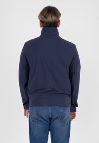 Giacca blu navy realizzata in tessuto morbido e liscio; presenta un colletto alto e polsini elasticizzati. Abbinata a jeans in denim azzurro chiaro.