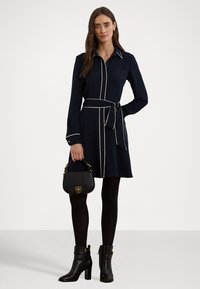 Vestido camisa azul-marinho com bainhas brancas, ajustado na cintura, combinado com botas de salto pretas e uma pequena bolsa preta com detalhes em dourado.