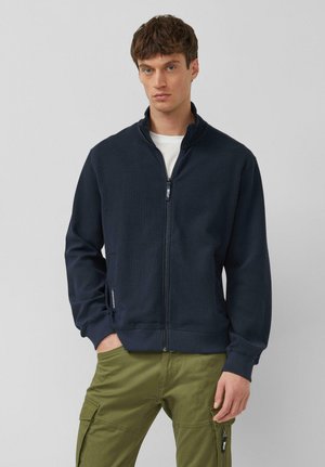 Man draagt een marineblauwe ritsjas over een wit overhemd met olijfgroene cargobroek, staand tegen een effen lichtgrijze achtergrond.