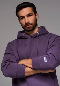 Haut à capuche violet avec une coupe décontractée, capuche à cordon de serrage et poignets côtelés. Présente une petite patch logo sur la manche. Texture lisse.