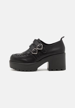 Zapato plataforma negro con tacón grueso, hebillas plateadas en forma de corazón, doble correa y costuras decorativas en la parte delantera.