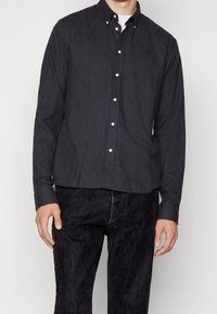 Camisa abotonada gris oscuro hecha de una tela texturizada, con cuello, botones dorados y un pequeño logo bordado en el pecho.