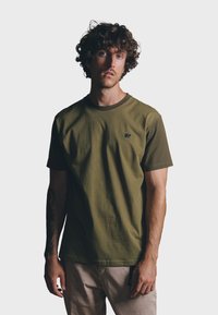 Camiseta de manga corta verde oliva con cuello oscuro contrastante y un pequeño logo de elefante en el lado izquierdo del pecho. Hecha de algodón suave.