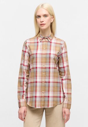 Donna con capelli biondi lisci che indossa una camicia a scacchi beige, bianca, rossa e nera, e pantaloni beige, in piedi contro uno sfondo semplice.