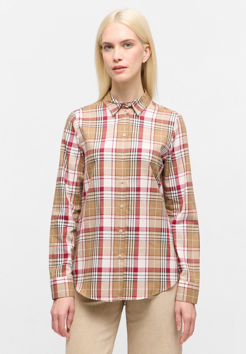 Woman with straight blonde hair wearing a beige, white, red, and black plaid button-up shirt and beige pants, standing against a plain background.