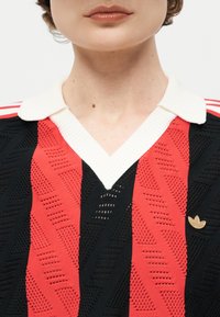 Άτομο που φοράει κόκκινο και μαύρο υφασμάτινο πλεκτό πουλόβερ με λευκό γιακά και ένα μικρό χρυσό καρφίτσα λογότυπο Adidas στη αριστερή πλευρά.