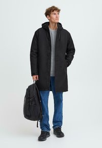 Črna kapuca parka z zadrgo, oblečena preko sivo puloverja, v kombinaciji z modrimi jeans hlačami in črnimi čevlji; drži črn nahrbtnik.