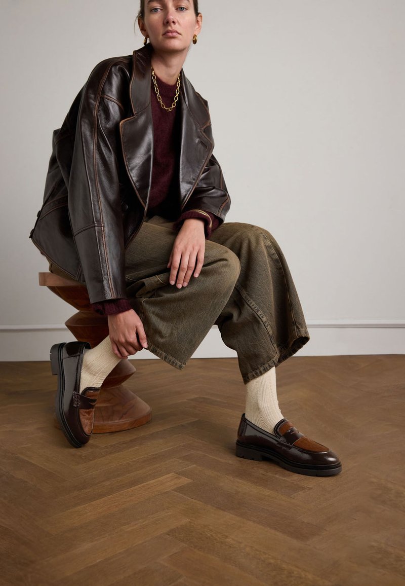 Braune Lederloafers mit strukturierten braunen Akzenten, kombiniert mit cremefarbenen Socken, olivgrünen weiten Hosen und einer dunkelbraunen oversized Jacke.