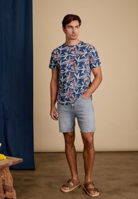 Marineblauwe t-shirt met kleurrijke bladerenpatronen, korte mouwen en een logo. Gecombineerd met lichte denim shorts en bruine sandalen.