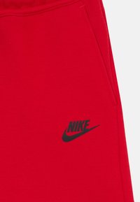 Sarkanas sporta bikses, kas izgatavotas no kokvilnas un sintētisko šķiedru maisījuma, ar melnu Nike logotipu kreisajā augšstilbā un sānu kabatu. Gluda tekstūra, elastīga josta.