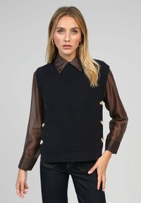 Débardeur en tricot sans manches de couleur bleu marine foncé avec un tissu texturé, présentant un col rond et des boutons dorés sur le côté. Associé à une chemise à col marron.