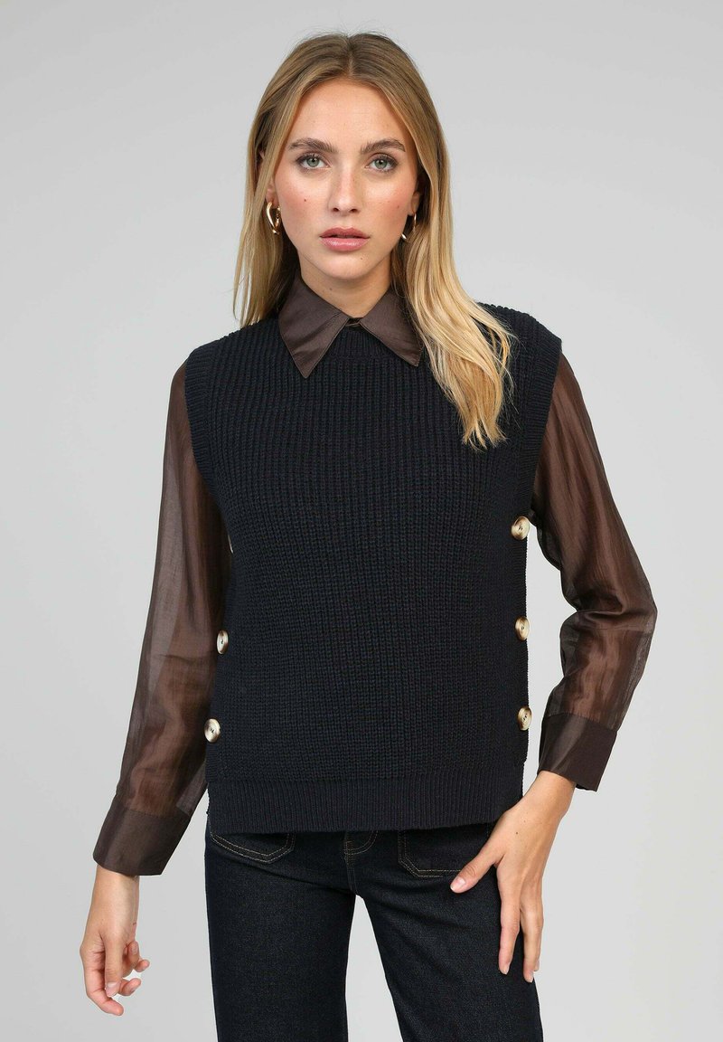 Débardeur en tricot sans manches de couleur bleu marine foncé avec un tissu texturé, présentant un col rond et des boutons dorés sur le côté. Associé à une chemise à col marron.