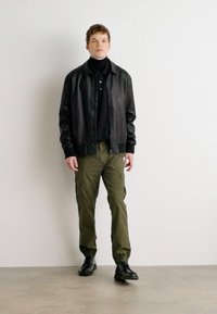 Jaqueta bomber de couro preta sobre um suéter preto de gola alta, combinada com calças cargo verde-oliva e sapatos pretos. Design simples, sem padrões visíveis.