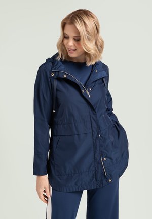 Femme portant une veste à capuche bleu marine avec fermeture éclair et boutons-pression, regardant vers le bas, les mains dans les poches, sur un fond clair uni.