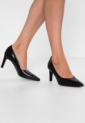 Jambes portant des talons aiguilles noirs texturés à bout pointu avec talons moyens sur fond blanc uni.