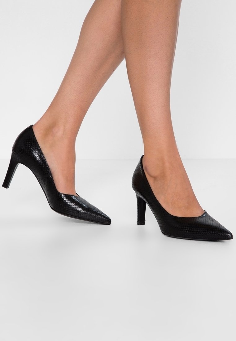 Jambes portant des talons aiguilles noirs texturés à bout pointu avec talons moyens sur fond blanc uni.