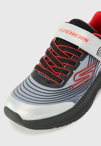 Baskets de sport grises avec accents rouges, lacets noirs et une sangle. Présente une tige texturée, un design incurvé et une semelle en caoutchouc.