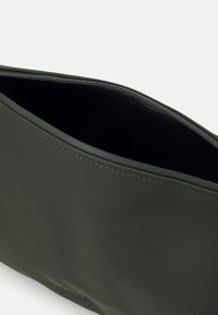 Borsetta verde oliva con una superficie liscia e opaca e foderata internamente di nero. Caratterizzata da una chiusura a zip e da un logo discreto sul lato.