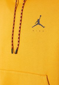Sudadera amarilla hecha de una tela suave. Presenta un logo negro de Jordan y cordones con un patrón que incluye toques de rojo, amarillo y negro. Incluye un bolsillo frontal.
