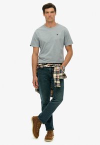 Grå kortærmet t-shirt, blå jeans, brune snørebåndsstøvler og en ternet skjorte bundet om taljen. Simpelt design, afslappet look.
