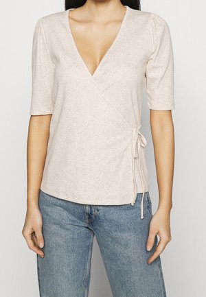 Vaaleanbeige kietoutuva paita, jossa on syvä V-kaula-aukko, lyhyet hihat ja sivusolkiyksityiskohta. Valmistettu pehmeästä, teksturoidusta kankaasta. Yhdistetty sinisiin farkkuihin.