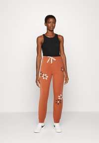 Calças de fato na cor ferrugem com padrão floral em branco e preto, cintura com cordão, punhos ajustados, combinadas com um top preto sem mangas e ténis brancos.