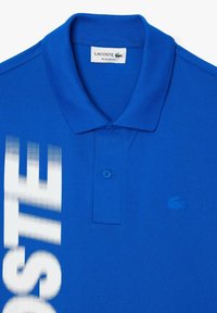 Sininen polo-paita, jossa on tekstuuri, kaksi nappia kauluksessa ja valkoinen graafinen logo. Omistaa hienovaraisen Lacosten krokotiilin embleemin.