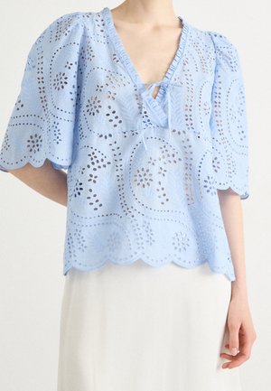 Bluzka z ażurowej koronki w jasnoniebieskim kolorze z falistymi krawędziami, krótkimi bufkami i wiązaniem przy dekolcie w kształcie litery V, noszona z białą spódnicą.