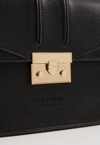 Primo piano di una borsa in pelle nera texture con chiusura rettangolare dorata e il logo in rilievo "SEIDENFELT MANUFAKTUR" sotto.