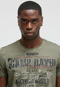 Olive green t-shirt made of cotton; features graphic text and distressed print, round neckline, and short sleeves.