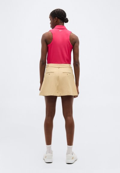 Kvinde med mørkt fletning hår iført en ærmeløs lyserød polo top, beige nederdel med baglommer, hvide sokker og hvide sneakers, stående med ryggen mod kameraet.