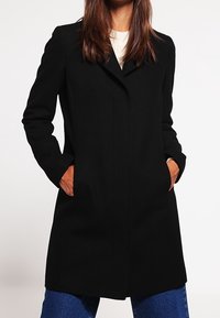 Svart kappa med skräddarsydd design, långa ärmar och sidofickor. Tillverkad av ett strukturerat tyg, med en skarp krage och dold fram.