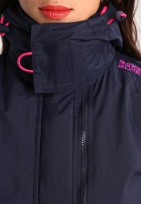 Navyblå vandtæt jakke med høj krave, lyserøde detaljer og lynlås foran. Har justerbare snoretræk ved halsen for en sikker pasform.