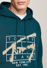 Sudadera color teal hecha de tela suave, con un diseño de logo blanco y un acento en aerosol beige. Incluye capucha con cordón y bolsillo frontal.