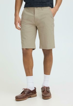 Muž oblečený v béžových šortkách dĺžky po kolená, bielych členkových ponožkách a hnedých kožených lodných topánkach stojí s rukami v vreckách.