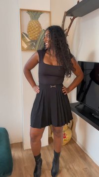 Schwarzes, tailliertes Oberteil mit quadratischem Ausschnitt, plissierter schwarzer Rock, goldene Schnallen-Details an der Taille, kombiniert mit schwarzen Knöchel-Stiefeln, drinnen fotografiert.