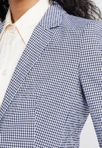 Femme portant un blazer à carreaux bleu et blanc sur une chemise crème boutonnée, avec des cheveux noirs bouclés visibles à l'arrière.