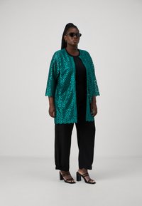 Teal kanten kimono jacket met bloemenpatroon over een zwarte top en zwarte broek, aangevuld met zwarte sandaaltjes met bandjes en een zonnebril.