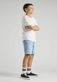 Biała koszulka z krótkim rękawem, jasnoniebieskie szorty dżinsowe, czarne sneakersy z białymi sznurówkami oraz skarpetki w czarne paski. Poza stojąca.